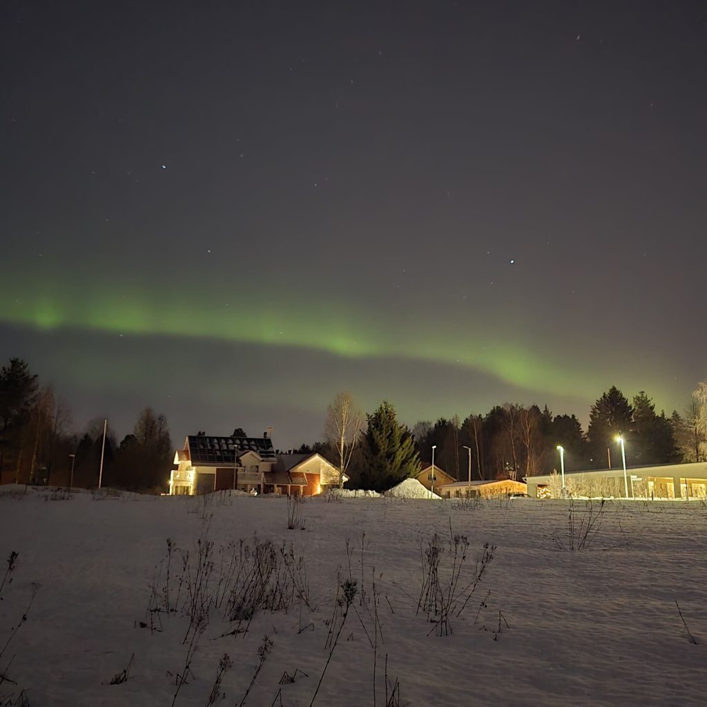 Une aurore boréale visible dans un quartier résidentiel de Rovaniemi, avec les lumières des maisons en dessous